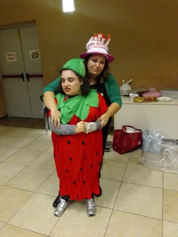 Una ragazza vestita da fragola, abbracciata da dietro da una volontaria con un cappello di festa.