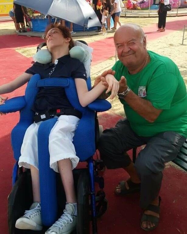 Un volontario tiene la mano di un ragazzo in carrozzina che, a occhi chiusi, ha il viso rivolto al sole.